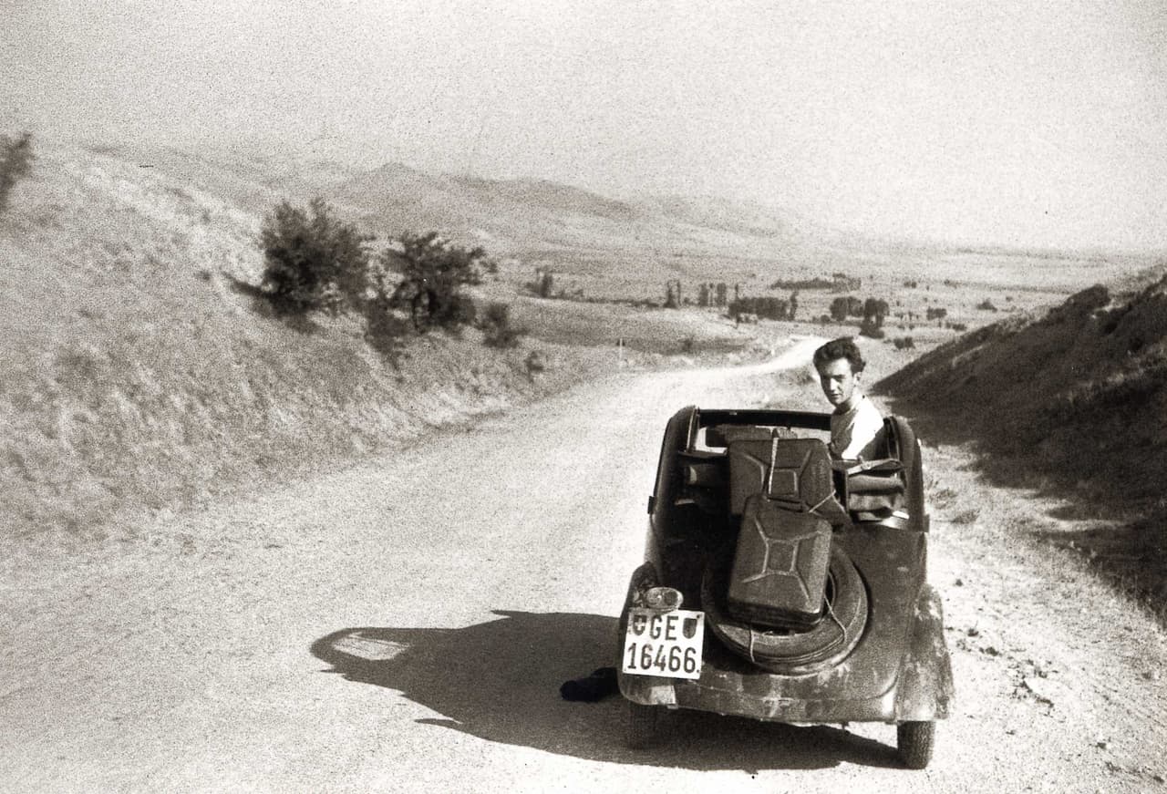 Photo d'archive en noir et blanc. La Fiat Topolino, immatriculée à Genève, sur la route d'Ankara. On voit la voiture de l'arrière, le capot est ouvert et Bouvier est retourné le siège passager, il regarde l'objectif. Deux valises sont attachée sur la roue de secours. La piste de gravillons s'étend jusqu'à l'horizon au pied de collines arides.